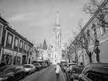 A black and white photograph features a narrow street lined with parked cars and bordered by buildings. In the distance, a prominent tower with intricate Gothic architectural details rises against the sky. A person wearing a long coat walks down the middle of the street, adding a sense of scale to the urban scene. Leafless trees line the sidewalks, contributing to a stark and atmospheric composition.