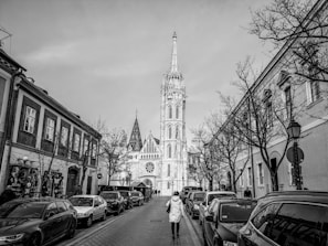 A black and white photograph features a narrow street lined with parked cars and bordered by buildings. In the distance, a prominent tower with intricate Gothic architectural details rises against the sky. A person wearing a long coat walks down the middle of the street, adding a sense of scale to the urban scene. Leafless trees line the sidewalks, contributing to a stark and atmospheric composition.