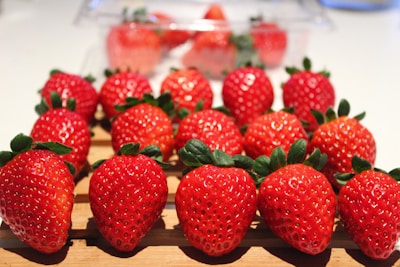 Bright, fresh strawberries are neatly arranged on a wooden surface with a few more strawberries in a clear plastic container in the background. The vibrant red fruit is contrasted by the dark green leaves, and the light reflects off their glossy skin.