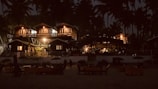 A nighttime scene featuring a series of cozy, illuminated wooden cabins surrounded by tall palm trees. The cabins have warm lights glowing, creating a welcoming atmosphere. In the foreground, there are several tables and umbrellas, suggesting a beach or resort setting.