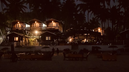 A nighttime scene featuring a series of cozy, illuminated wooden cabins surrounded by tall palm trees. The cabins have warm lights glowing, creating a welcoming atmosphere. In the foreground, there are several tables and umbrellas, suggesting a beach or resort setting.
