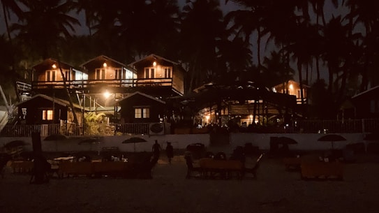 A nighttime scene featuring a series of cozy, illuminated wooden cabins surrounded by tall palm trees. The cabins have warm lights glowing, creating a welcoming atmosphere. In the foreground, there are several tables and umbrellas, suggesting a beach or resort setting.