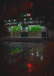 A pharmacy storefront is illuminated against the dark night, with green signage and a striped awning. The wet pavement in front reflects colorful lights, including red and green hues, creating a vivid, almost cinematic appearance. Abstract shapes and signs are visible on the store's glass doors.