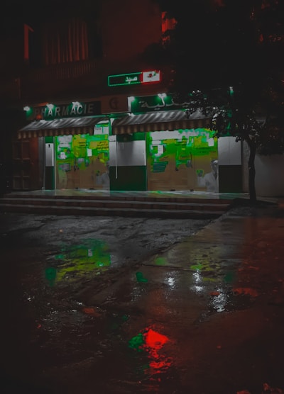 A pharmacy storefront is illuminated against the dark night, with green signage and a striped awning. The wet pavement in front reflects colorful lights, including red and green hues, creating a vivid, almost cinematic appearance. Abstract shapes and signs are visible on the store's glass doors.