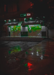 A pharmacy storefront is illuminated against the dark night, with green signage and a striped awning. The wet pavement in front reflects colorful lights, including red and green hues, creating a vivid, almost cinematic appearance. Abstract shapes and signs are visible on the store's glass doors.