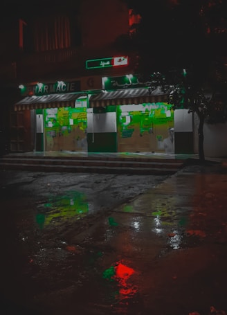 A pharmacy storefront is illuminated against the dark night, with green signage and a striped awning. The wet pavement in front reflects colorful lights, including red and green hues, creating a vivid, almost cinematic appearance. Abstract shapes and signs are visible on the store's glass doors.