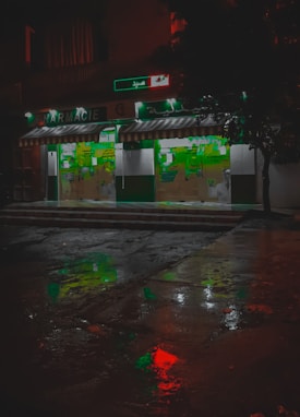 A pharmacy storefront is illuminated against the dark night, with green signage and a striped awning. The wet pavement in front reflects colorful lights, including red and green hues, creating a vivid, almost cinematic appearance. Abstract shapes and signs are visible on the store's glass doors.