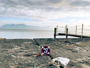 A close-up view of a bottle cap with the letters 'RES KP' resting on a weathered stone surface. In the background, a serene body of water stretches towards distant mountains under a partly cloudy sky. A wooden pier extends over the water, lined with simple rope railings.