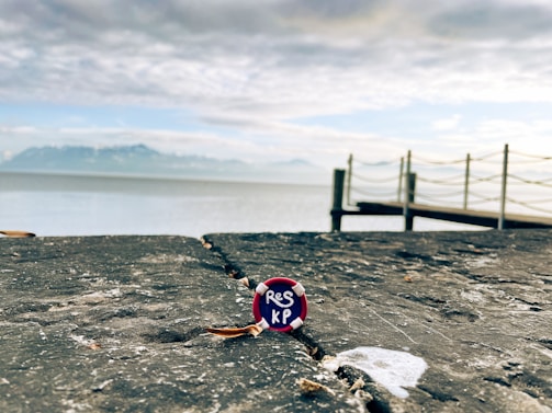 A close-up view of a bottle cap with the letters 'RES KP' resting on a weathered stone surface. In the background, a serene body of water stretches towards distant mountains under a partly cloudy sky. A wooden pier extends over the water, lined with simple rope railings.