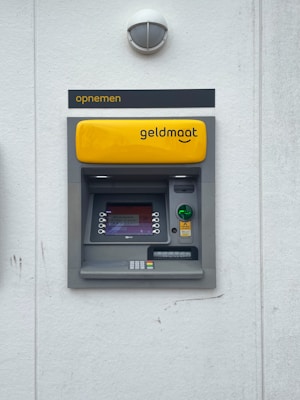 A modern ATM embedded in a wall, featuring a bright yellow panel with the word 'geldmaat' on it. The machine includes a screen displaying text, a numeric keypad, and card slots. Above the ATM, there's a sign with the text 'opnemen'. The background is a light gray wall with a small, round light fixture on top.