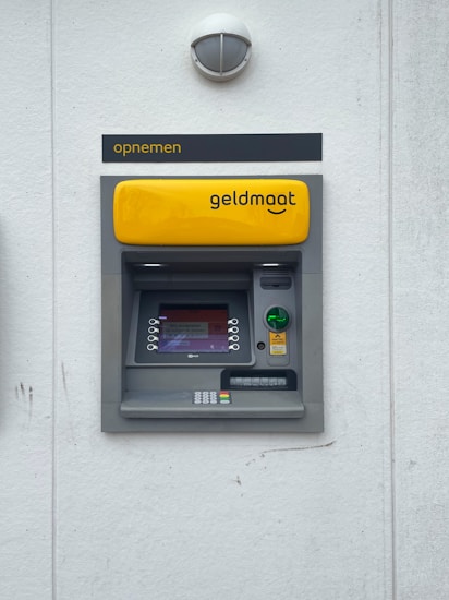 A modern ATM embedded in a wall, featuring a bright yellow panel with the word 'geldmaat' on it. The machine includes a screen displaying text, a numeric keypad, and card slots. Above the ATM, there's a sign with the text 'opnemen'. The background is a light gray wall with a small, round light fixture on top.