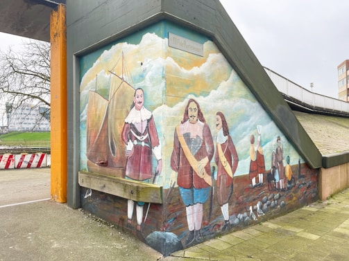 A mural painted on a concrete structure depicts historical figures in period clothing, including men with long coats, sashes, and boots. The scene includes a sailing ship, seagulls, and a group of people by a rocky shore. The background features a cloudy sky and distant buildings.