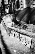 A series of paintings are displayed on the ground along the steps of a building. The artworks vary in style and subject, and the setting appears to be an outdoor walkway with fallen leaves scattered on the brick pavement. Shadows cast by surrounding trees and structures create an interplay of light and dark on the scene.