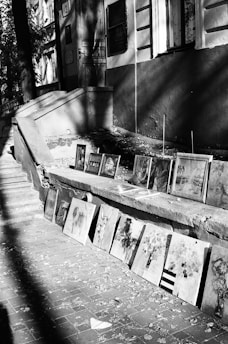 A series of paintings are displayed on the ground along the steps of a building. The artworks vary in style and subject, and the setting appears to be an outdoor walkway with fallen leaves scattered on the brick pavement. Shadows cast by surrounding trees and structures create an interplay of light and dark on the scene.