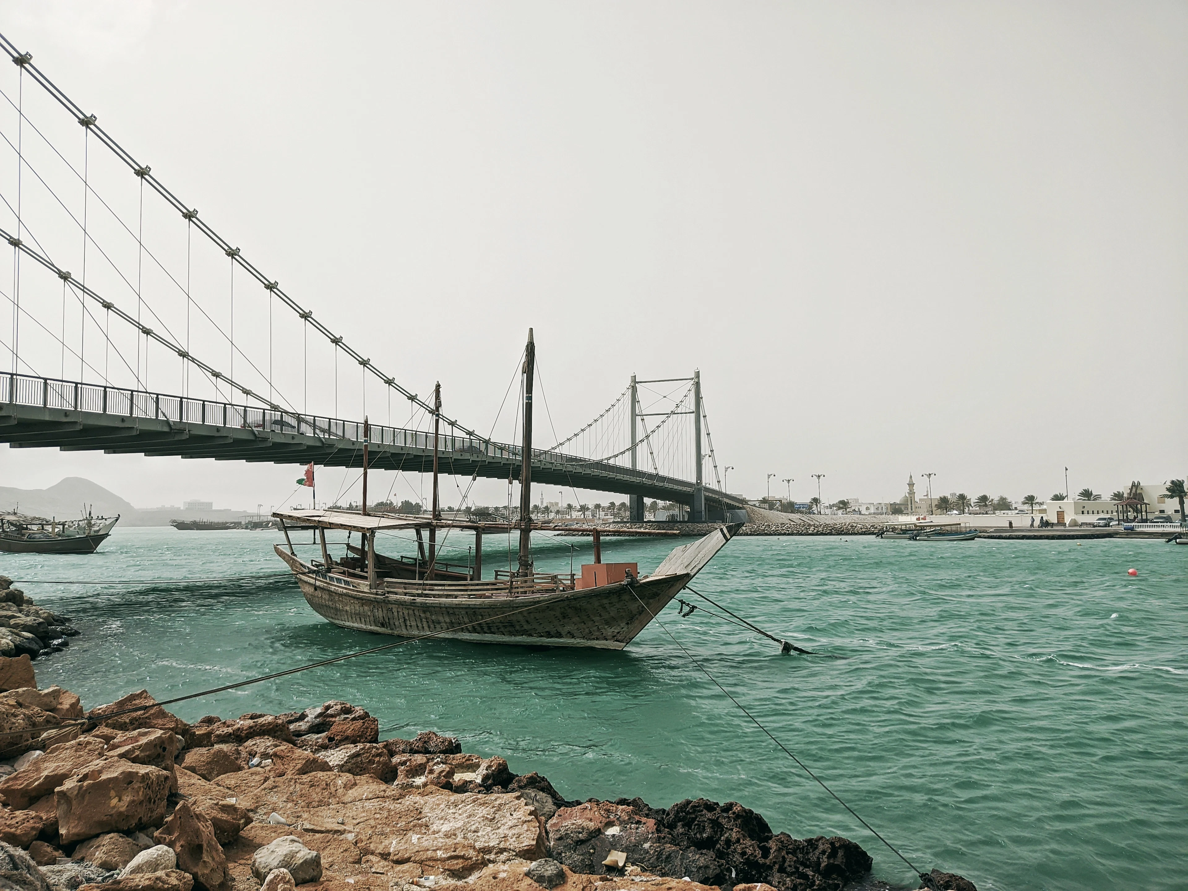 A wooden dhow moored in a turquoise inlet beneath a suspension bridge, Oman