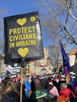 A large group of people gathered outdoors, holding a sign that reads 'Protect Civilians in Ukraine' with yellow hearts. Flags and signs with various colors and slogans are visible among the crowd. Trees without leaves and buildings are present in the background under a clear blue sky.