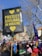 A large group of people gathered outdoors, holding a sign that reads 'Protect Civilians in Ukraine' with yellow hearts. Flags and signs with various colors and slogans are visible among the crowd. Trees without leaves and buildings are present in the background under a clear blue sky.