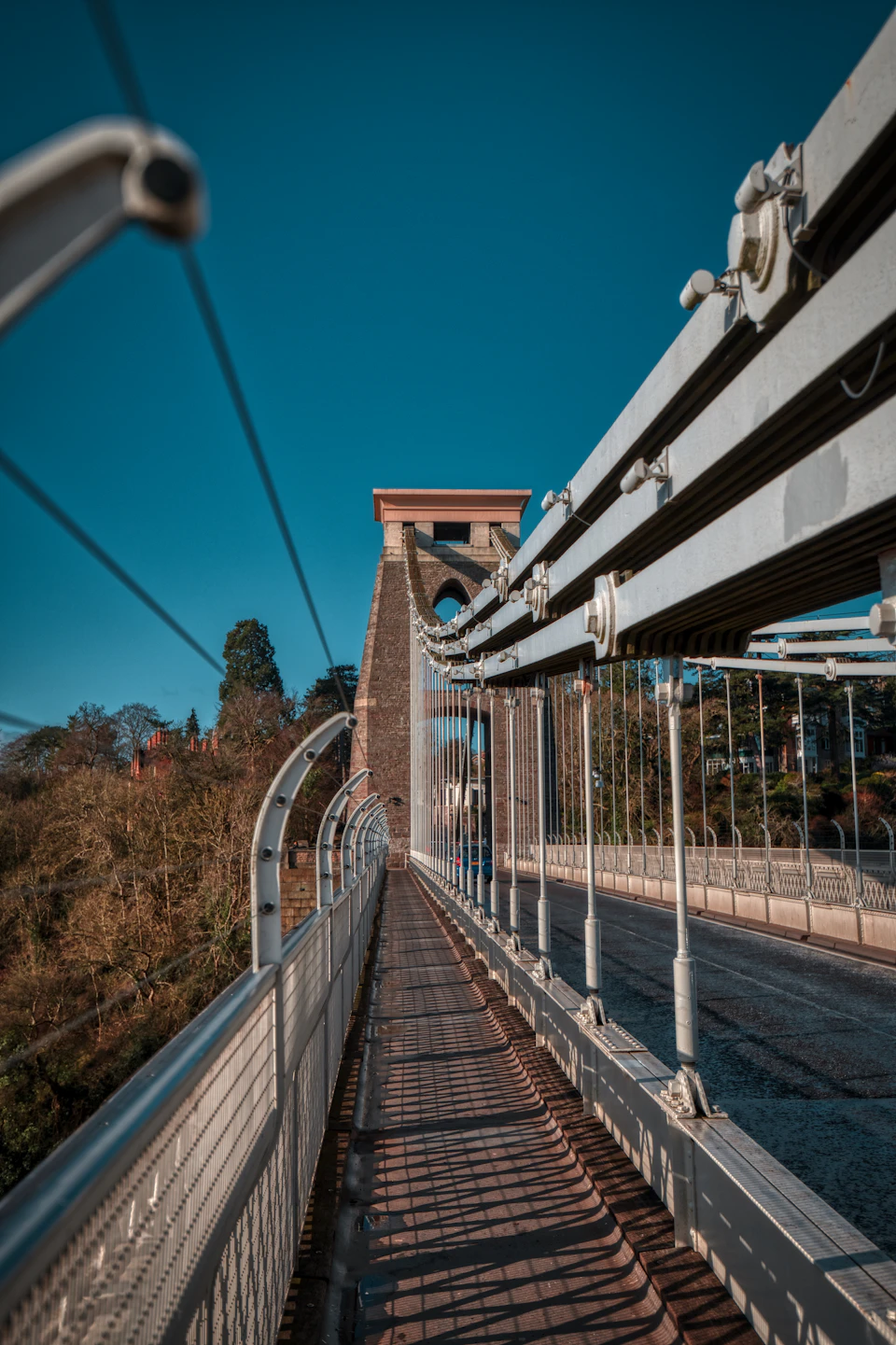 The image shows a suspension bridge with multiple cables and wires, painted white, stretching across a river or valley. The bridge is supported by stone pillars that add to its architectural charm. A pedestrian walkway runs along the length of the bridge, providing access for pedestrians. The sky above is clear blue, creating a beautiful contrast with the white structure below. In the distance, trees can be seen on either side of the river or valley, adding a touch of nature to the scene.