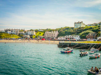A scenic seaside village with a sandy beach where people are swimming and enjoying the sun. Colorful houses and buildings are nestled on lush green hills overlooking the water. Several small boats with red buoys are anchored near a stone pier, and the calm sea reflects the sky's blue hue.