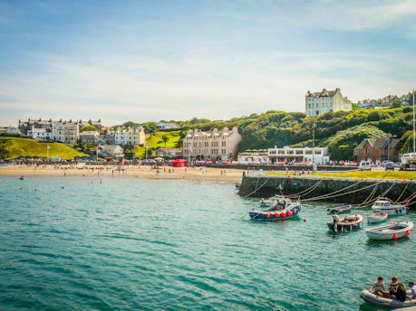 A scenic seaside village with a sandy beach where people are swimming and enjoying the sun. Colorful houses and buildings are nestled on lush green hills overlooking the water. Several small boats with red buoys are anchored near a stone pier, and the calm sea reflects the sky's blue hue.