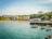 A scenic seaside village with a sandy beach where people are swimming and enjoying the sun. Colorful houses and buildings are nestled on lush green hills overlooking the water. Several small boats with red buoys are anchored near a stone pier, and the calm sea reflects the sky's blue hue.