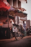 A bicycle is positioned in front of a street vendor stall, equipped with a large metallic container on its rear rack. The stall is shaded by colorful umbrellas, and various goods are visible, including bags and baskets. The setting appears to be a bustling outdoor market with warm, natural lighting.