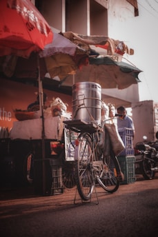 A bicycle is positioned in front of a street vendor stall, equipped with a large metallic container on its rear rack. The stall is shaded by colorful umbrellas, and various goods are visible, including bags and baskets. The setting appears to be a bustling outdoor market with warm, natural lighting.