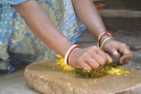 Hands adorned with red and golden bangles are grinding a yellow substance on a stone slab using a stone pestle. The person is wearing a blue and yellow floral patterned dress.