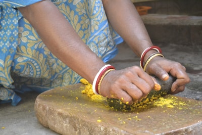 Hands adorned with red and golden bangles are grinding a yellow substance on a stone slab using a stone pestle. The person is wearing a blue and yellow floral patterned dress.