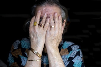 An elderly person is covering their face with both hands. The hands are adorned with rings, bracelets, and the person is wearing a patterned garment.