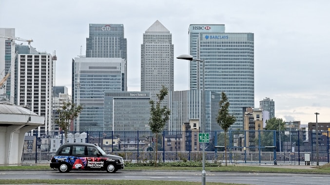A cityscape featuring tall modern skyscrapers representing major financial institutions like Citibank, HSBC, and Barclays. In the foreground, a black taxi with an advertisement is parked on the street. The sky appears overcast and the surrounding area includes some trees and street lamps.