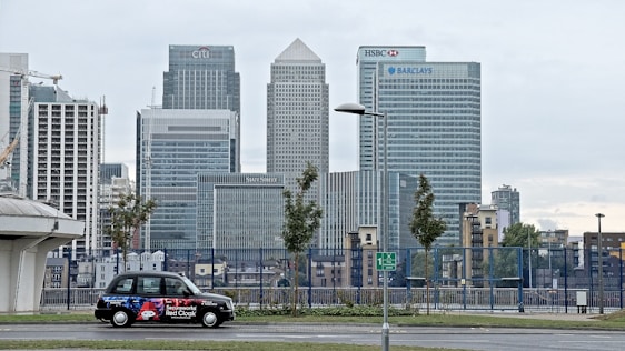 A cityscape featuring tall modern skyscrapers representing major financial institutions like Citibank, HSBC, and Barclays. In the foreground, a black taxi with an advertisement is parked on the street. The sky appears overcast and the surrounding area includes some trees and street lamps.