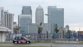 A cityscape featuring tall modern skyscrapers representing major financial institutions like Citibank, HSBC, and Barclays. In the foreground, a black taxi with an advertisement is parked on the street. The sky appears overcast and the surrounding area includes some trees and street lamps.