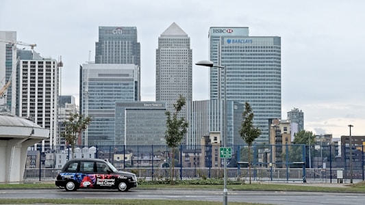 A cityscape featuring tall modern skyscrapers representing major financial institutions like Citibank, HSBC, and Barclays. In the foreground, a black taxi with an advertisement is parked on the street. The sky appears overcast and the surrounding area includes some trees and street lamps.