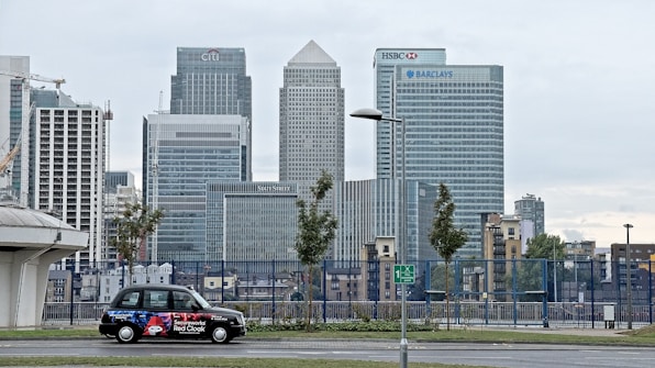 A cityscape featuring tall modern skyscrapers representing major financial institutions like Citibank, HSBC, and Barclays. In the foreground, a black taxi with an advertisement is parked on the street. The sky appears overcast and the surrounding area includes some trees and street lamps.