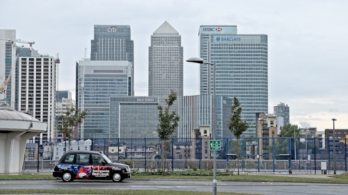A cityscape featuring tall modern skyscrapers representing major financial institutions like Citibank, HSBC, and Barclays. In the foreground, a black taxi with an advertisement is parked on the street. The sky appears overcast and the surrounding area includes some trees and street lamps.