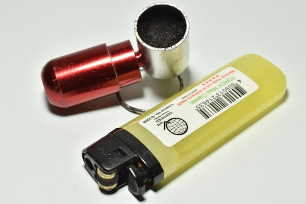 A yellow lighter is lying on a white surface with its bottom facing upwards, revealing a barcode label. Next to the lighter, there is a small metallic container with a red cap and a black substance inside.