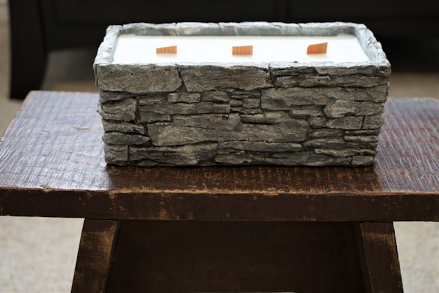 A rectangular stone-textured container filled with white material, possibly wax, positioned on a dark wooden table. Three wooden wicks are visible emerging from the wax-like substance.