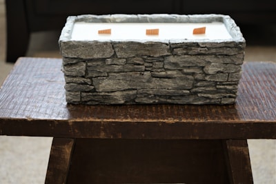 A rectangular stone-textured container filled with white material, possibly wax, positioned on a dark wooden table. Three wooden wicks are visible emerging from the wax-like substance.
