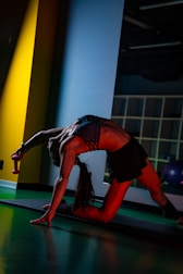 A person is engaged in a yoga or fitness pose on a mat, illuminated by colored lighting. They are holding a small dumbbell and their body is stretched gracefully, showcasing strength and flexibility. The environment suggests an indoor gym or studio setting, with vibrant blue, red, and yellow lights creating a dynamic and energetic atmosphere.