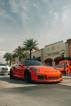 A bright orange sports car is prominently parked on a sunny street lined with palm trees and boutique shops. Nearby, a white luxury car and another vehicle with an open hood can be seen. The atmosphere appears stylish and leisurely, with clear skies overhead.