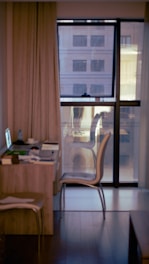 A small workspace is set up next to a large window in a contemporary interior. A desk holds a laptop, papers, and a cup, suggesting the area is used for work or study. Two chairs are placed around the desk. The window spans from floor to ceiling, offering a blurred view of an urban apartment building outside. Neutral-colored curtains partially cover the window, and the room appears to have a cozy, modern ambiance.