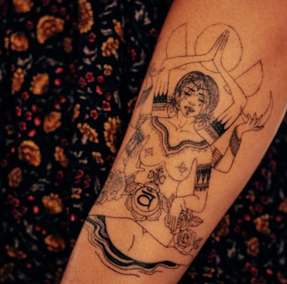 A detailed tattoo on a person's skin depicts a woman with multiple arms in a meditative or spiritual pose. The design incorporates intricate patterns, flowers, and spiritual symbols.