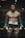A shirtless man stands confidently in a boxing ring, wearing black shorts with flame designs and boxing gloves. He has a tattoo on his thigh, and his hair is tied back. The setting is dimly lit, creating a moody and intense atmosphere.