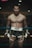 A shirtless man stands confidently in a boxing ring, wearing black shorts with flame designs and boxing gloves. He has a tattoo on his thigh, and his hair is tied back. The setting is dimly lit, creating a moody and intense atmosphere.