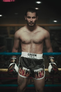A shirtless man stands confidently in a boxing ring, wearing black shorts with flame designs and boxing gloves. He has a tattoo on his thigh, and his hair is tied back. The setting is dimly lit, creating a moody and intense atmosphere.