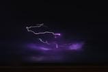 A dark sky is illuminated by bright, jagged streaks of purple lightning. The atmosphere is stormy and intense, with deep shadows and a sense of movement in the clouds.