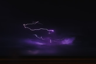 A dark sky is illuminated by bright, jagged streaks of purple lightning. The atmosphere is stormy and intense, with deep shadows and a sense of movement in the clouds.
