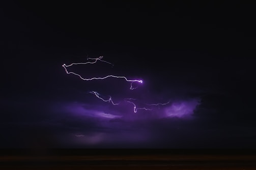 A dark sky is illuminated by bright, jagged streaks of purple lightning. The atmosphere is stormy and intense, with deep shadows and a sense of movement in the clouds.