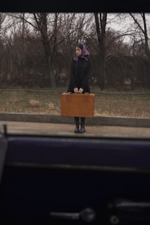 A woman stands on the roadside near barren trees, holding a large brown suitcase. She wears a black coat, boots, and a purple headscarf with white polka dots. The scene is observed from inside a vehicle.